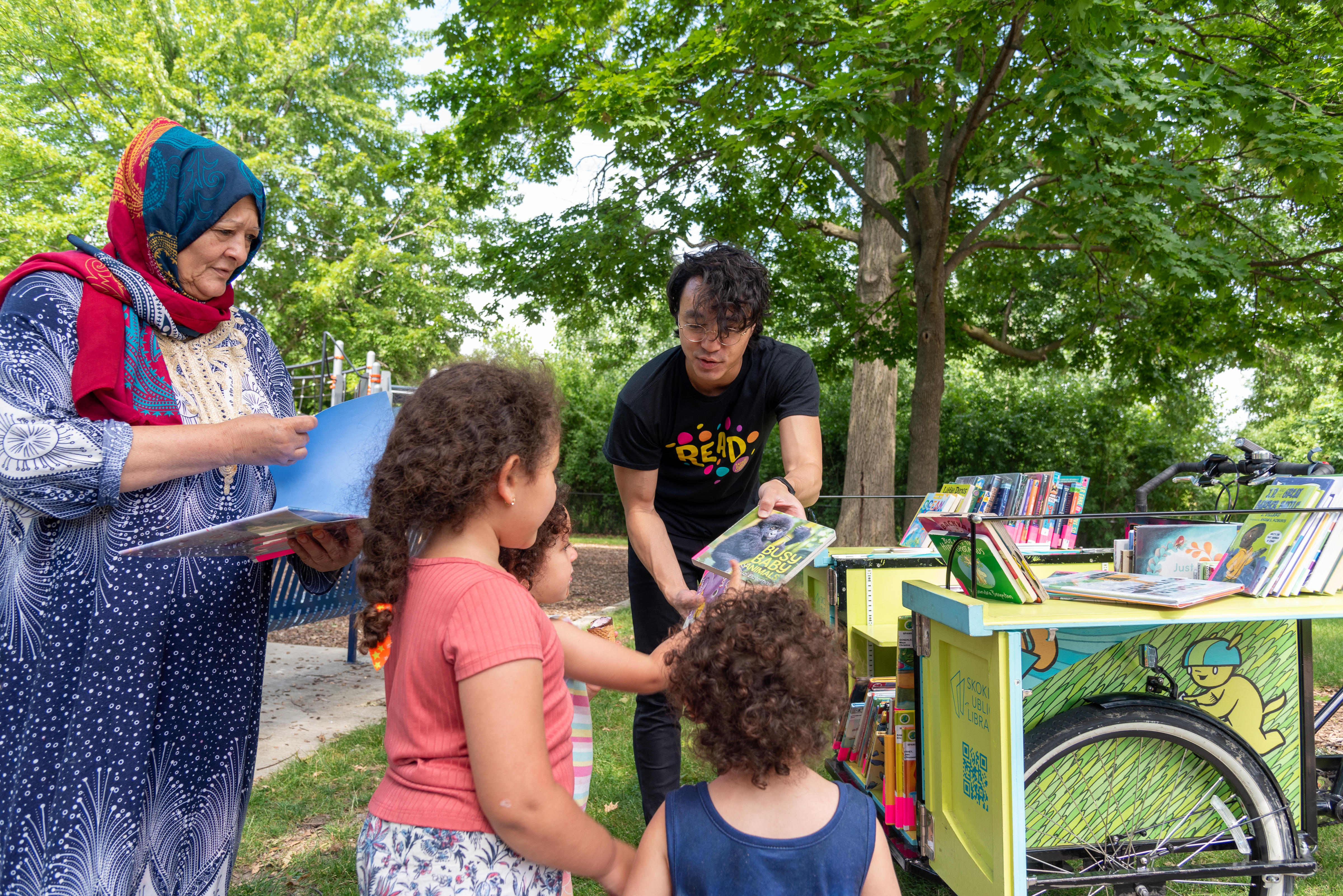 A library staff member is showing a picture book to three children. An adult is standing next to them looking at another picture book. 