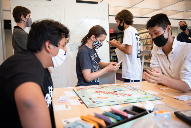 Three teen patrons in the foreground are playing Monopoly while another teen patron does schoolwork on a whiteboard with a staff member. Another staff member can be seen in the very back of the photo.