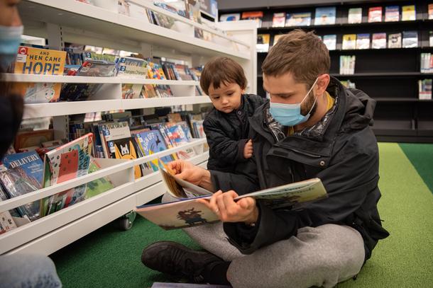 A father sits cross legged on the green striped floor reading a book to his small son, who is not even as tall as the man sitting down. The boy holds onto his father's black jacket.