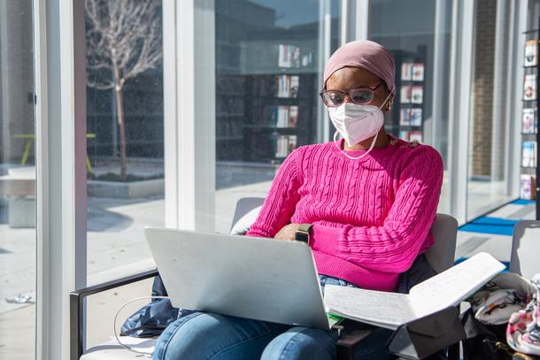 A masked woman wearing a bright pink sweater sits in a chair with a computer and an open notebook on her lap. There is light coming in from a window on her right, which overlooks an outdoor courtyard.