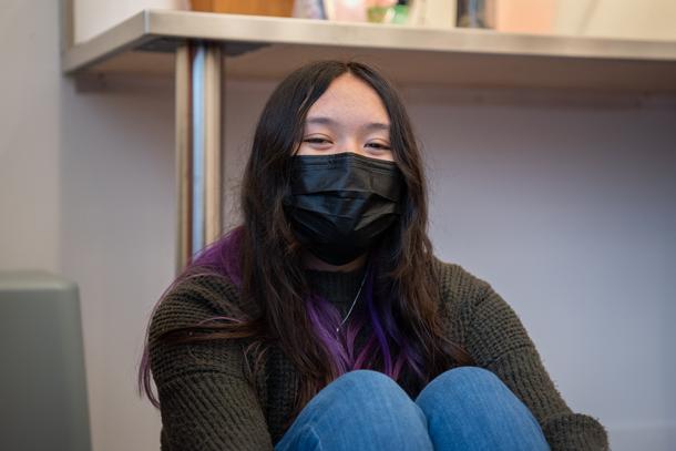 A teenager with long brown hair with purple streaks sits on the floor with her knees to her chest. She is wearing a mask.