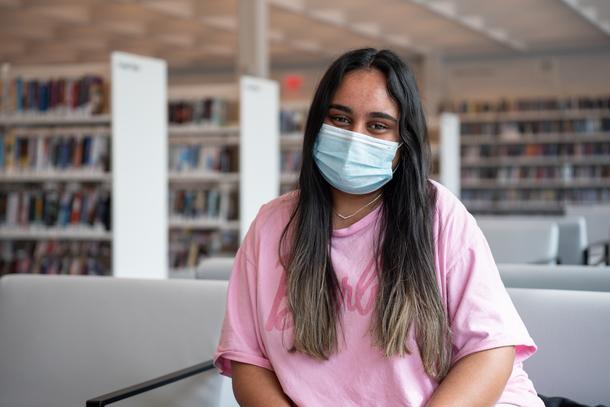 A young woman with long brown hair, a mask, and a pink t-shirt sits in a chair with shelves of books behind her.