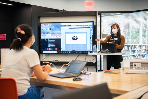 A staff member in a brown top and black apron gestures to a digital display showing a program for making vinyl cut stickers. There is a lotus sticker on the screen, and a patron in a white top faces the display, listening.